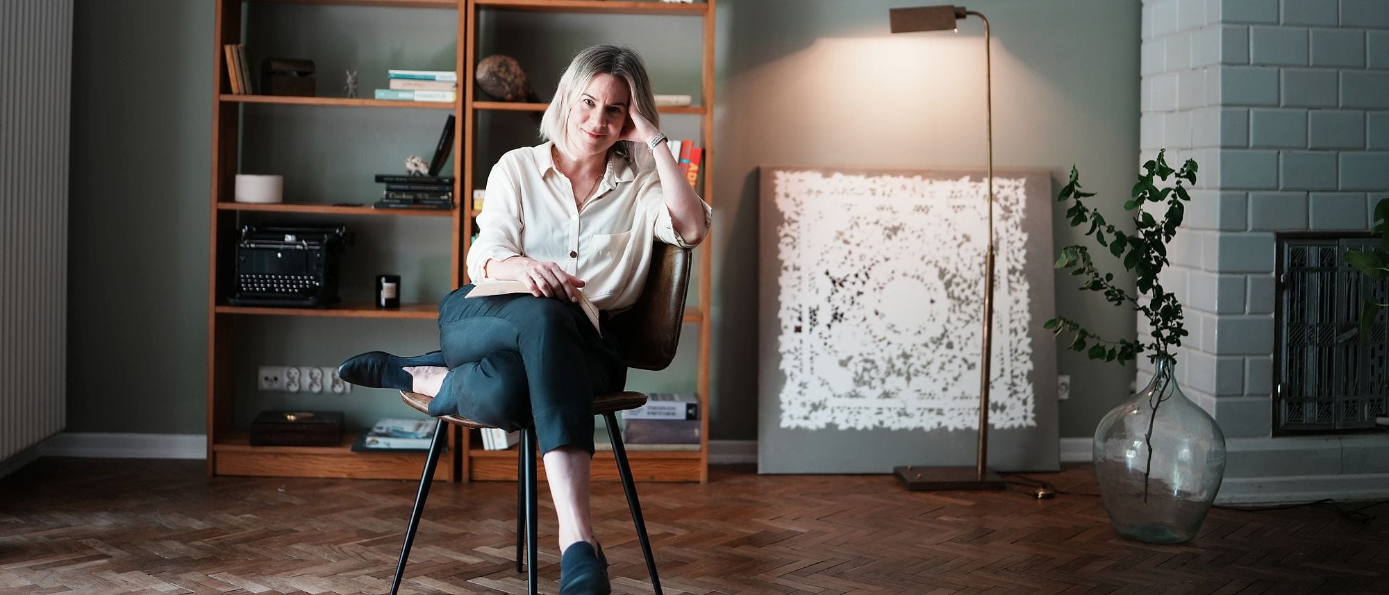 Szeroki plan gabinetu psychoterapii: Anna Wyganowska-Błażejewska na krześle przy regale z książkami, obok lampa i kaflowy piec