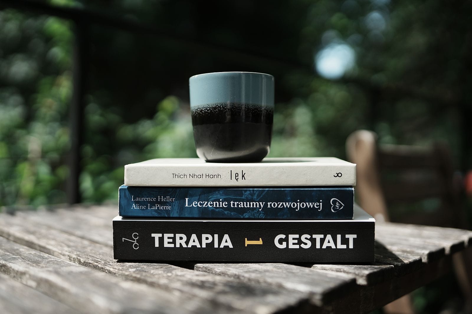 Stos książek: „Lęk”, „Leczenie traumy rozwojowej” i „Terapia Gestalt”, na nich kubek, w tle zieleń