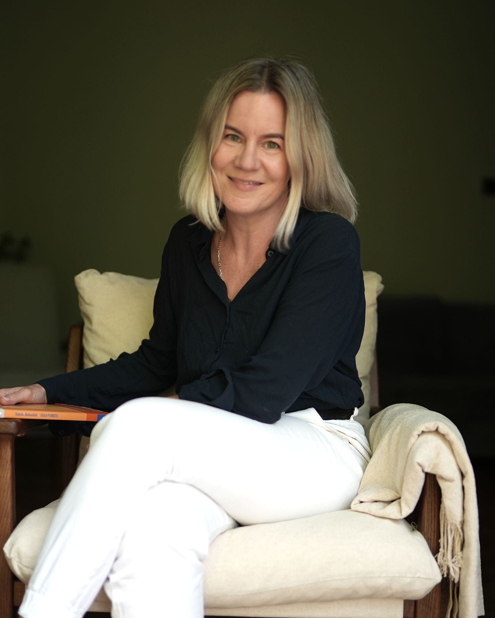 Anna Wyganowska-Błażejewska, psychoterapeutka Gestalt, siedzi w fotelu w gabinecie z książką w dłoni i uśmiecha się