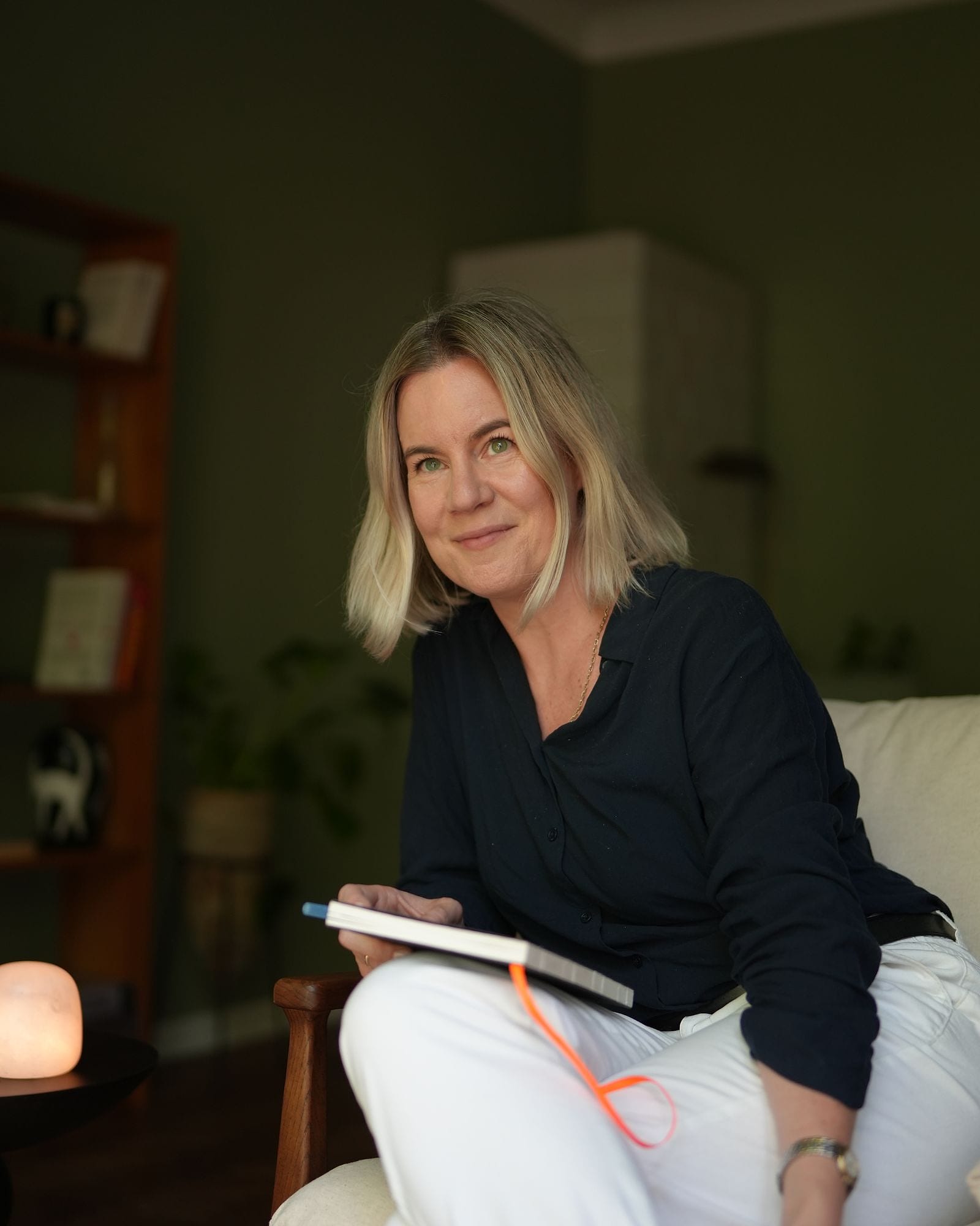 Anna Wyganowska-Błażejewska z notesem w fotelu, obok zapalona lampa solna i regał z książkami