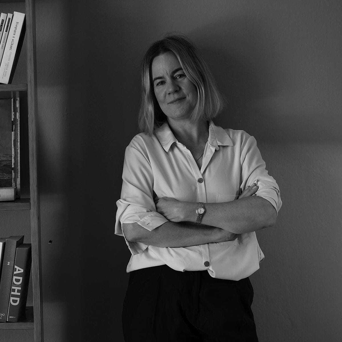 Anna Wyganowska-Błażejewska stoi ze skrzyżowanymi rękami przy regale z książkami w gabinecie, zdjęcie czarno-białe
